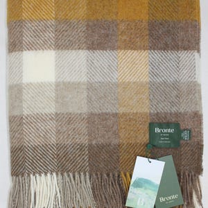 Könnte beinhalten: Eine gefaltete Decke mit einem Karomuster in Gold-, Braun- und Cremetönen. Die Decke hat einen Fransenrand und ein Etikett mit der Aufschrift "Woodale Gold Pure New Wool".