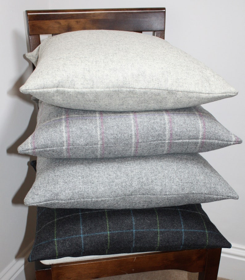 May include: Four square decorative pillows stacked on a wooden chair. The top pillow is a solid light gray, followed by a gray pillow with a pink and white plaid pattern. The third pillow is a solid gray, and the bottom pillow has a dark gray plaid pattern with blue and green accents.