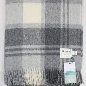 Può includere: Coperta piegata in lana merino grigia Port Ellen con motivo a quadri nei toni del grigio e crema. La coperta ha un bordo con frange e un'etichetta con la scritta "Bronte by Moon".