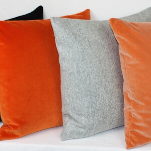 May include: Four decorative throw pillows in a variety of colors and textures. The pillows are arranged in a row, with a black velvet pillow on the left, an orange velvet pillow in the middle, a gray wool pillow in the center, and another orange velvet pillow on the right.