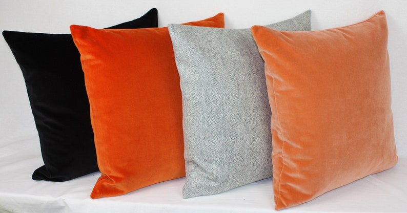May include: Four decorative throw pillows in a variety of colors and textures. The pillows are arranged in a row, with a black velvet pillow on the left, an orange velvet pillow in the middle, a gray wool pillow in the center, and another orange velvet pillow on the right.