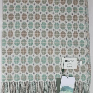 Peut inclure: Une couverture pliée avec un motif floral répété vert menthe et marron sur fond blanc. La couverture a une frange sur le bord inférieur. L'étiquette indique "Milan Mint Merino Wool".