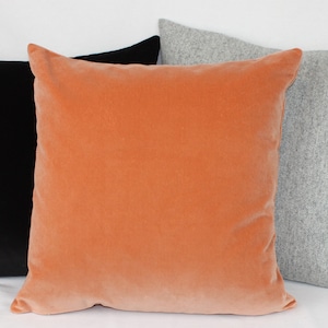 May include: Three decorative throw pillows in different colors and textures. The pillow on the left is black velvet, the middle pillow is a peachy orange velvet, and the pillow on the right is a light gray wool.