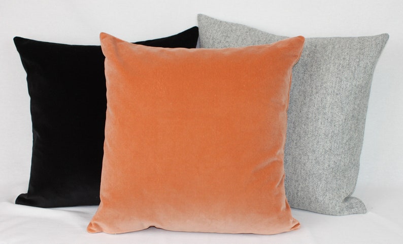 May include: Three decorative throw pillows in different colors and textures. The pillow on the left is black velvet, the middle pillow is a peachy orange velvet, and the pillow on the right is a light gray wool.