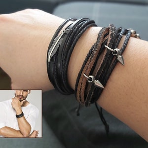 May include: A set of layered bracelets with black and brown leather cords, metal accents, and a feather charm.