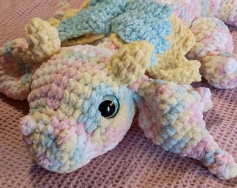 Peut inclure: Un dragon en crochet fait à la main aux couleurs pastel rose, jaune et bleu. Le jouet en peluche a de grands yeux brillants et un aspect doux et texturé. Le dragon est posé sur une couverture rose.