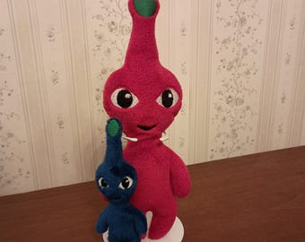 May include: A plush toy set featuring a large magenta creature with a green sprout on its head and a smaller blue creature with a green sprout on its head. Both figures have large white eyes with black pupils and stand on a circular white base.