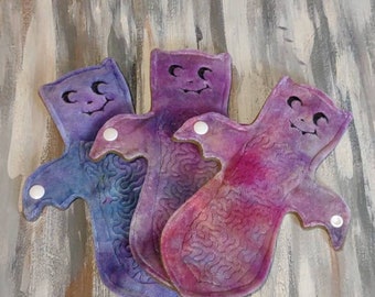 May include: Three reusable cloth menstrual pads in shades of purple and blue, each with a smiling face design. The pads have a curved shape with wings and snap closures. The background is a textured, abstract painting.