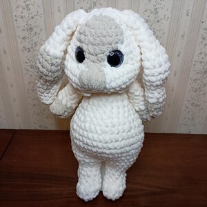 May include: A handmade, plush bunny toy in white and gray. The bunny has large, black eyes with blue highlights, long floppy ears, and a fluffy, textured body. The toy is standing upright, showcasing its soft, cuddly appearance.