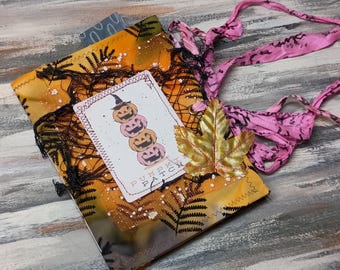 Peut inclure: Un journal fait à la main sur le thème d'Halloween avec une illustration de citrouilles empilées sur la couverture. Le journal présente un tissu orange, jaune et noir avec des motifs de fougères et de toiles d'araignées. Un ruban rose et une feuille d'or ajoutent des accents décoratifs.