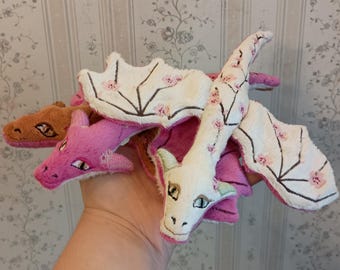 May include: A plush toy featuring three dragon heads in brown, pink, and white. The white dragon head and wings are decorated with small pink cherry blossom patterns. The toy has embroidered eyes and detailed wing structures with dark outlines.
