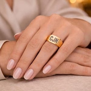 Può includere: Un anello con sigillo dorato con le iniziali "CJN" incise in nero. L'anello ha una faccia rettangolare e viene indossato su un dito. Lo sfondo è sfocato, con un focus sull'anello e sulla mano.