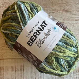 Könnte beinhalten: Ein Knäuel Bernat Blanket-Garn in Grün-, Blau- und Grautönen. Das Garn ist dick und weich mit einer weichen Textur. Auf dem Etikett steht "Bernat Blanket" und "Yarnspirations".