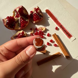May include: A still life featuring a halved pomegranate with bright red seeds, a miniature hazelnut, cinnamon sticks, and the word "Pomegranate" written on a white strip. The scene is set on a white surface.