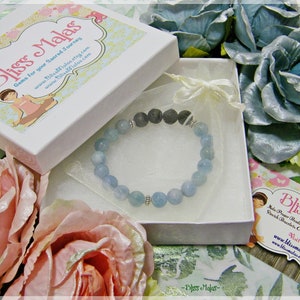 Puede incluir: Un brazalete de piedra de aguamarina azul claro con una cuenta de lava negra y separadores plateados. El brazalete está en una caja de regalo blanca con un forro de satén blanco. La caja está rodeada de flores artificiales rosas y azules.