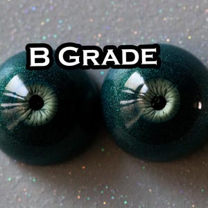 May include: Two domed, circular cabochons resembling realistic eyes with deep forest green, glitter-infused irises and pale, textured pupils. The text B GRADE is printed in white with a black outline centered above the two eye-shaped pieces.