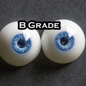 May include: A pair of round, white artificial eyes with detailed blue irises and dark pupils. The text B GRADE is printed in white with a black outline between the two eyes. The eyes have a smooth, glossy finish and a realistic appearance.