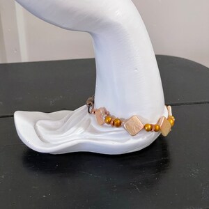 May include: A bracelet featuring square, iridescent, light brown beads and small, round, gold-colored beads. The bracelet is displayed on a white, sculpted hand-shaped form, set against a dark background.