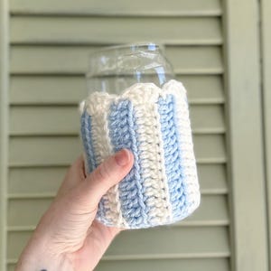 May include: A crocheted beverage insulator with blue and white stripes. The insulator is made of yarn and is designed to fit around a glass or can.
