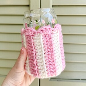 May include: A pink and white crocheted cup cozy with a scalloped top. The cozy is on a glass cup with a floral design.