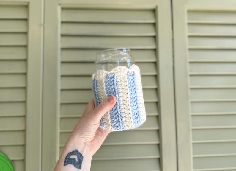 May include: A crocheted beverage insulator with blue and white stripes. The insulator is made of yarn and is designed to fit around a glass or can.