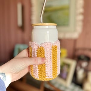 May include: A white ceramic travel mug with a wooden lid and a pink and yellow crocheted cozy. The mug has a stainless steel straw sticking out of the lid.
