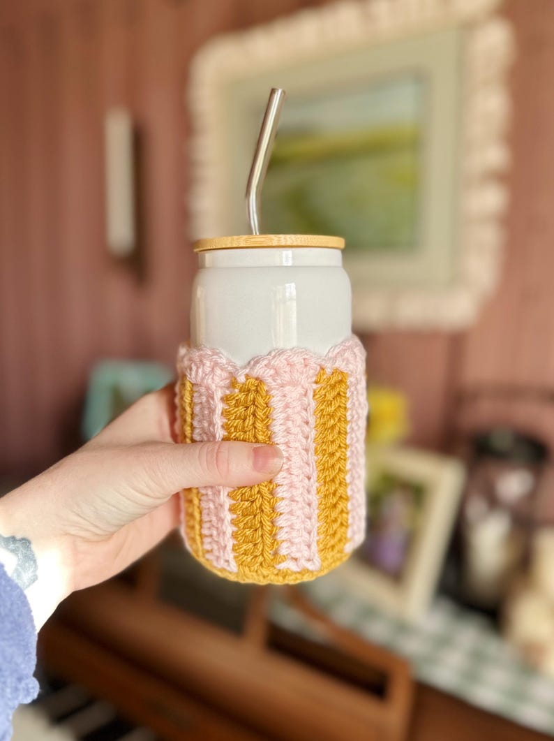 May include: A white ceramic travel mug with a wooden lid and a pink and yellow crocheted cozy. The mug has a stainless steel straw sticking out of the lid.