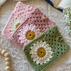 May include: Three crocheted phone cases with daisy designs. The cases are pink, green, and a darker shade of pink. Each case has a white daisy with a yellow center.