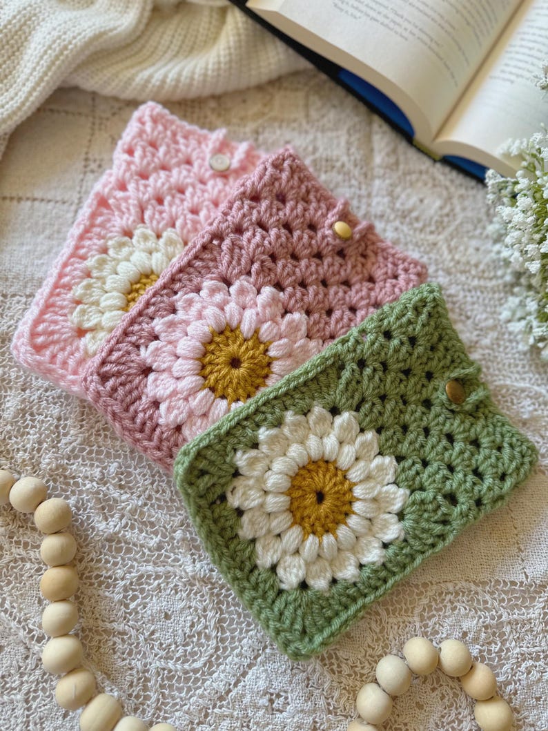 May include: Three crocheted phone cases with daisy designs. The cases are pink, green, and a darker shade of pink. Each case has a white daisy with a yellow center.