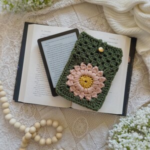 May include: A green crocheted e-reader case with a pink and yellow flower design. The case is lying on top of an open book with a white background.