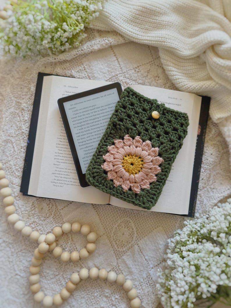 May include: A green crocheted e-reader case with a pink and yellow flower design. The case is lying on top of an open book with a white background.