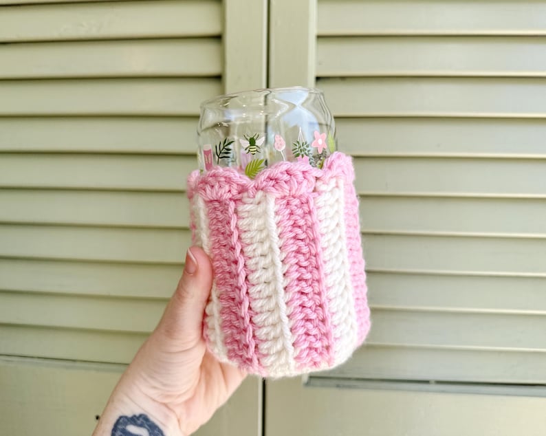 May include: A pink and white crocheted cup cozy with a scalloped top. The cozy is on a glass cup with a floral design.