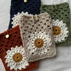 May include: Three crocheted phone cases with a daisy design. The cases are in shades of brown, beige, and green.