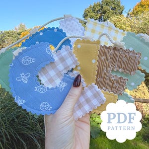 May include: A fabric bunting banner featuring scalloped-edge letters in various colors and patterns, including blue with ladybugs and bees, yellow gingham, and brown wood grain prints, all strung together on a thick white rope cord.