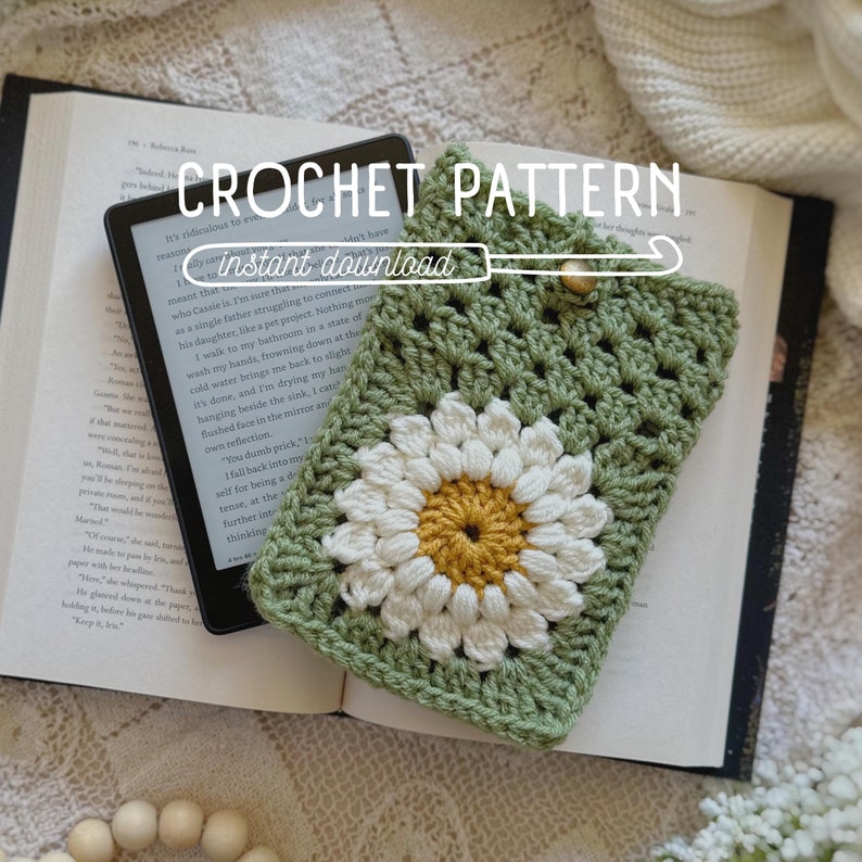 May include: Green crochet phone case with a white daisy flower design. The case is lying on a book with the text "CROCHET PATTERN instant download" visible.