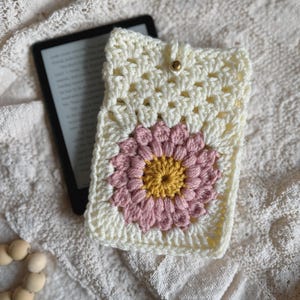 May include: A crocheted white book cover with a pink and yellow flower design. The cover has a button closure.
