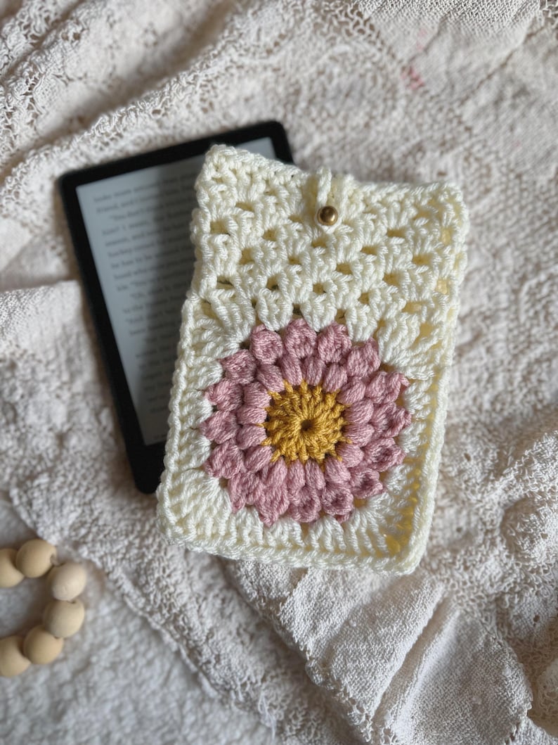 May include: A crocheted white book cover with a pink and yellow flower design. The cover has a button closure.