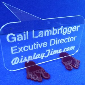 May include: A clear acrylic sign with a speech bubble shape and a white engraved message that reads "Gail Lambrigger Executive Director DisplayTime.com". The sign is propped up on two red plastic feet.