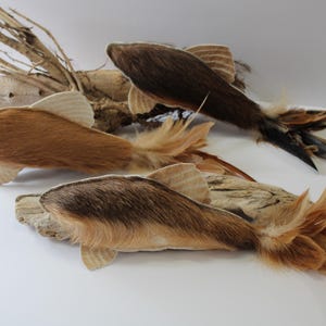 Peut inclure: Trois ornements en forme de poisson fabriqués à partir de fourrure brune et noire, avec des nageoires en tissu clair et des queues composées de plumes superposées. Les poissons sont placés près de morceaux de bois flotté clair et texturé.