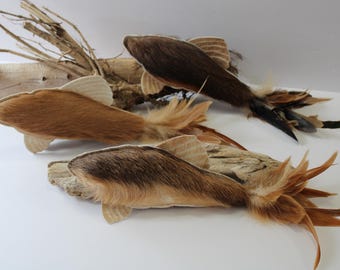 May include: Three fish-shaped ornaments crafted from brown and black fur with light-coloured fabric fins and tails made of layered feathers. The fish are positioned near pieces of light-coloured driftwood, showcasing textured fur and detailed feather accents.
