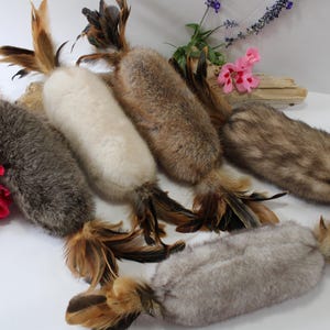 May include: A collection of five cylindrical cat toys made of soft, textured fur in shades of white, grey, and brown. Each toy is accented with long, dark brown and tan feathers attached to both ends, creating a distinct, elongated shape.