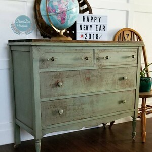 Pu&ograve; includere: Una credenza vintage verde con quattro cassetti e gambe tornite. La credenza ha una finitura invecchiata ed &egrave; decorata con un globo terrestre e un cartello che recita "Happy New Year 2018".