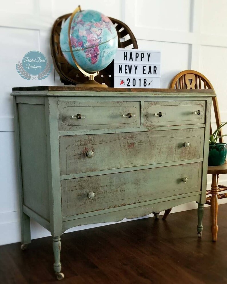 Pu&ograve; includere: Una credenza vintage verde con quattro cassetti e gambe tornite. La credenza ha una finitura invecchiata ed &egrave; decorata con un globo terrestre e un cartello che recita "Happy New Year 2018".