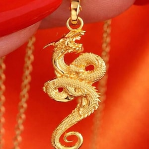 Puede incluir: Un colgante de dragón dorado con detalles intrincados, suspendido de una delicada cadena. El dragón está representado en una pose enrollada con la boca abierta. El fondo es de color naranja vibrante.