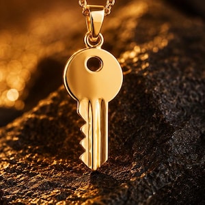 Puede incluir: Un collar con colgante de llave dorado. El colgante tiene forma de llave detallada con una parte superior circular y una paleta dentada. La cadena del collar también es dorada. El fondo es una superficie oscura y texturizada.