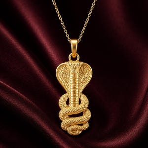 Pode incluir: Colar com pingente dourado com design de cobra. O pingente retrata uma cobra com o capuz levantado e o corpo enrolado. O colar tem uma corrente delicada e está sobre um fundo bordô.