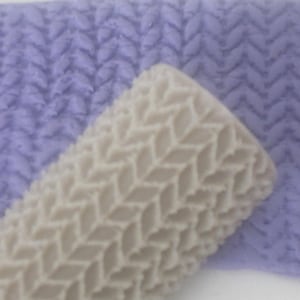 May include: A light beige rolling pin with a textured knit pattern is positioned in front of a lavender background with a matching knit pattern. The rolling pin is cylindrical and appears to be made of a hard material.