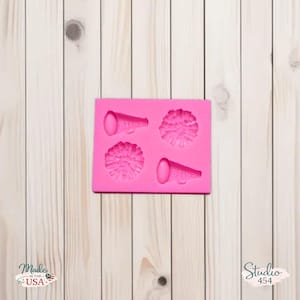 May include: A bright pink silicone mold featuring two pom-poms and two megaphones. The mold is rectangular and sits on a light wood-grain surface. The words "Made in the USA" and "Studio 454" are visible.