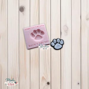 May include: A light pink square mould with a paw print design and the text "MADE IN THE USA" and "BAKER'S TOY BOX". A black and light blue paw print sticker is next to the mould. The Studio 454 logo is also visible.