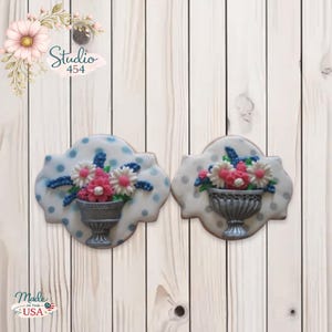 May include: Two decorated sugar cookies. Each cookie has a light blue and white polka dot background. A gray vase filled with pink and white flowers is the focal point. The cookies are in a decorative shape. The text "Studio 454" is visible.